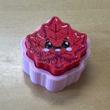 A small red glittery maple leaf-shaped clicker with a cute face, sitting in a lavender plastic mold or tray on a light wood surface.
