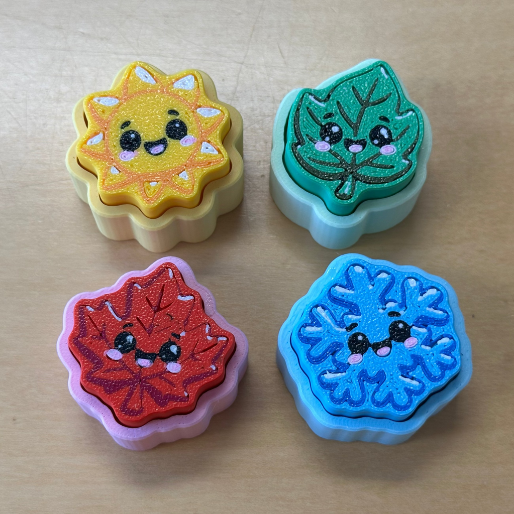 Four cute, kawaii-style autumn leaf-shaped clickers arranged in a 2x2 grid on a light wood surface. The leaves represent the four seasons, each in a different color: yellow sun, green leaf, red maple, and blue snowflake, all with embroidered smiling faces.