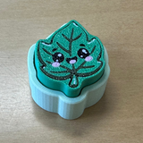 A small leaf-shaped clicker with a cute smiling face, resting on a white flower-shaped base on a light wood surface.