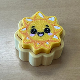 A small sun-shaped yellow clicker with a cute cartoon face, resting on a beige scalloped base on a light wood surface.