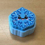 A small blue snowflake-shaped clicker with a cute embroidered face, placed on a light wood tabletop.